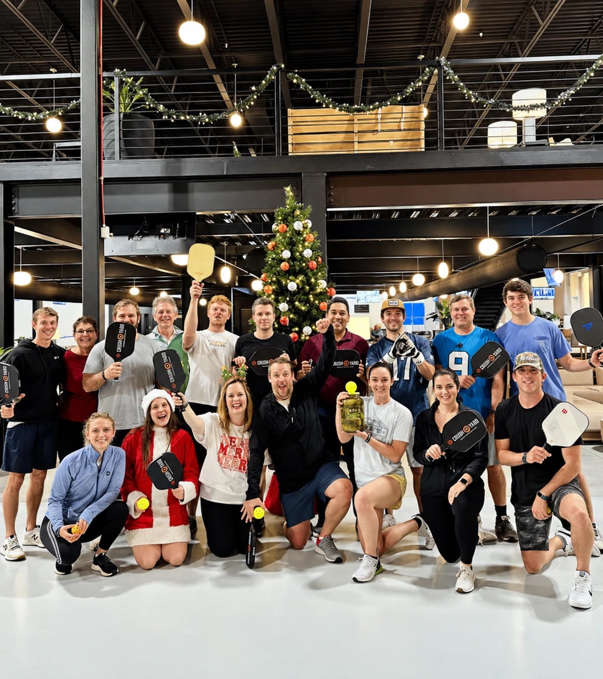 A holiday group celebrating with pickleball paddles beside a Christmas tree