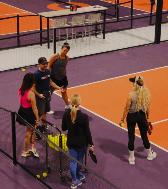 Five people stand on an indoor pickleball court, holding paddles and balls. A nearby bench holds bags and water bottles.