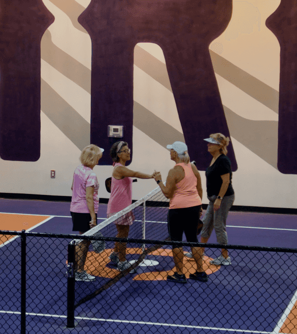 Four women on an indoor tennis court shake hands over the net. Two wear pink tops, two wear black. The court has a bold, colorful design.