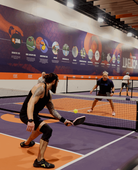 Two people play pickleball indoors on a court with vibrant graphics on the walls. One player swings at the ball near the net.