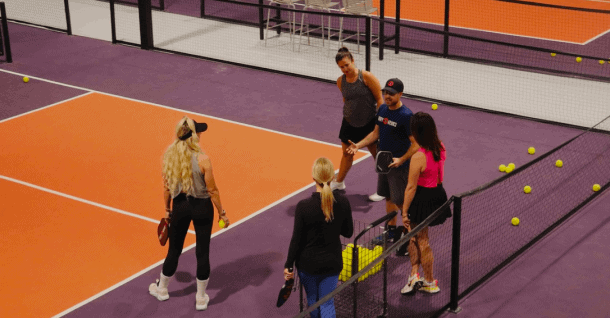 Four people stand on a purple and orange indoor court discussing, holding paddles, with tennis balls scattered around.