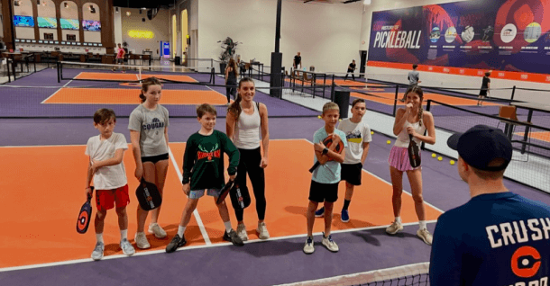 A group of children with paddles stand on an indoor pickleball court, listening to an instructor.