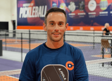 A man in a blue shirt holding a pickleball paddle stands in front of an indoor pickleball court.