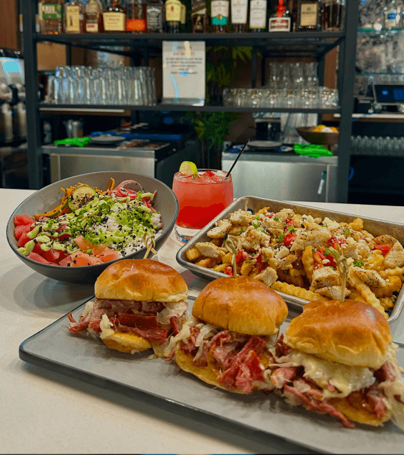 A bar counter with a salad, three sandwiches, loaded fries, and a pink cocktail with lime. Bottles and glasses are in the background.