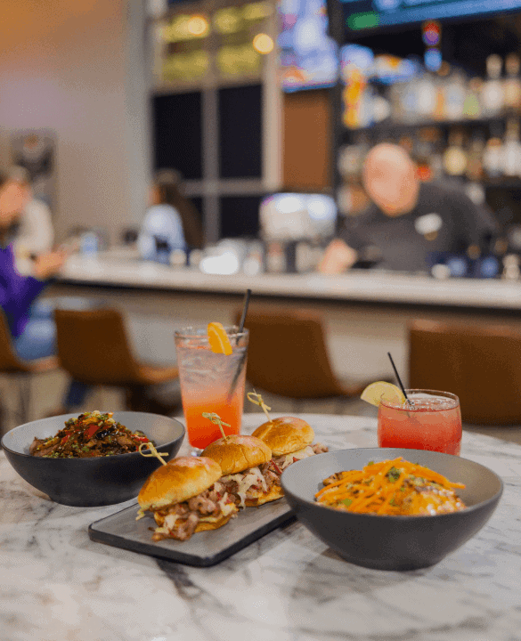 A restaurant table with sliders, pasta, and drinks. Bar counter and people in the background.