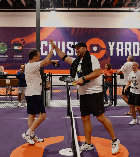 Two people on a purple indoor tennis court celebrate a point with a high-five. Spectators in the background watch the match.