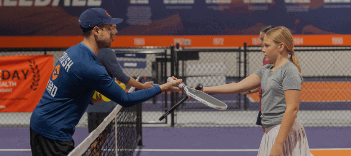 A tennis coach instructs a young player on a court, showing her how to hold a racket.