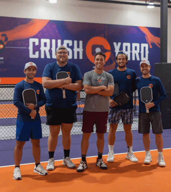 Five people standing on an indoor court, each holding a pickleball paddle. A large "Crush Yard" sign is visible in the background.