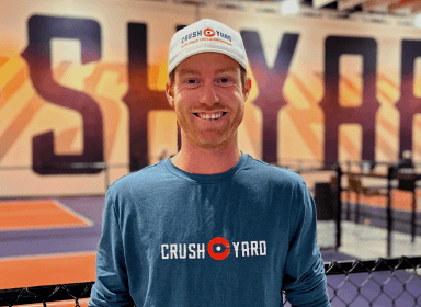 Smiling man wearing a Crush Yard shirt and cap stands in front of a large mural indoors.