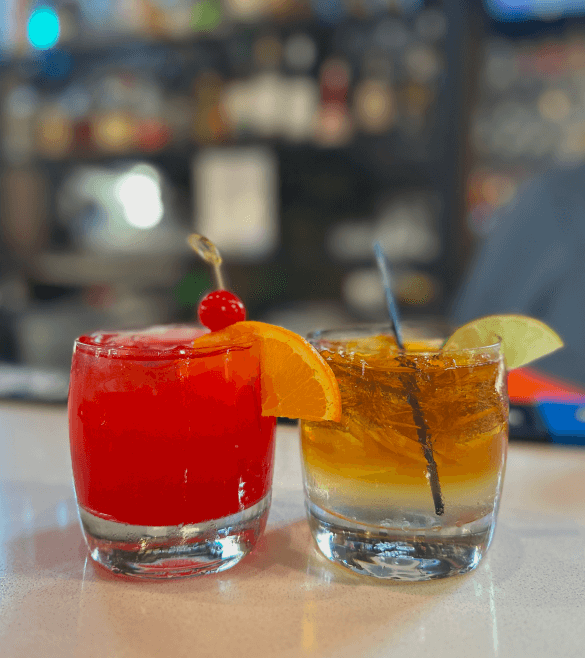 Two cocktails in short glasses on a bar counter; the left drink is red with an orange slice and cherry, while the right drink is amber with a lime wedge and stirrer.