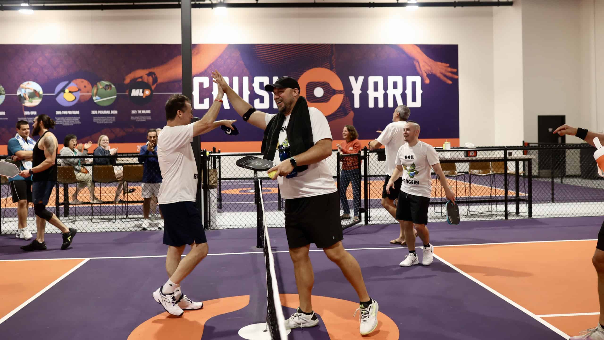 Two men high-five on an indoor pickleball court while others watch and play in the background at a sports venue called "Crush Yard.
